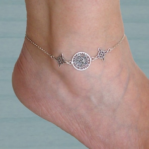 Può includere: Una cavigliera argentata con un pendente centrale circolare con un disegno a stella, affiancato da due ciondoli decorativi. La cavigliera è indossata su una caviglia, mostrando la delicata catena e gli intricati dettagli del gioiello.