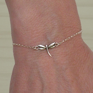 Op de afbeelding: Een delicate goudkleurige armband met een libellenbedel. De libel is gecentreerd op een fijne ketting, waarbij de armband op een pols rust. De libellenbedel heeft gedetailleerde vleugels en een gebogen staart.