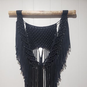 May include: Black macrame wall hanging with a decorative design, suspended from a natural wooden dowel. The macrame features a central white skull and long fringe. The piece is a unique, handcrafted home decor item.