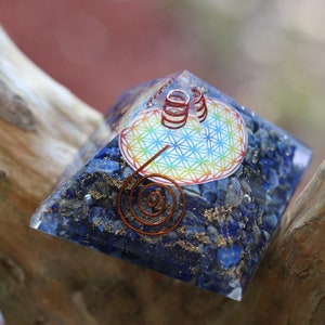 May include: A blue crystal pyramid with a copper wire design and a rainbow flower of life symbol. The pyramid is sitting on a wooden surface.