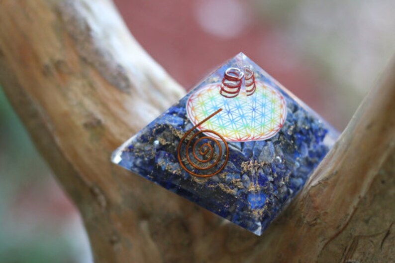 May include: A blue crystal pyramid with a copper wire spiral and a flower of life design. The pyramid is made of lapis lazuli and is sitting on a wooden surface.