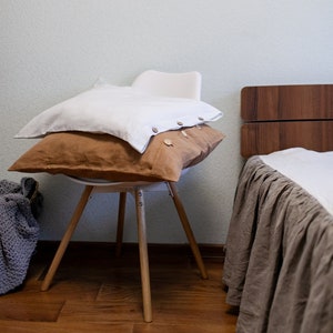 Może przedstawiać: Dwie poszewki lniane, jedna biała, a druga brązowa, leżą na białym krześle z drewnianymi nogami. Krzesło stoi przed łóżkiem z brązowym zagłówkiem i brązową lnianą spódnicą łóżka.