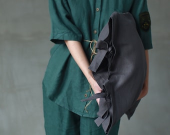 May include: A dark gray linen tote bag with multiple tie closures is held in front of a person wearing a green linen shirt and pants. The shirt has a button-down design and short sleeves. The background is a solid gray.