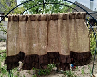 May include: A brown burlap curtain with a dark brown ruffled trim. The curtain is hanging on a black metal arch in a garden setting. The curtain is made of a woven material and has a rustic, farmhouse aesthetic.