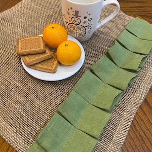 May include: A burlap placemat with a green ruffled edge. The placemat has a white plate with three biscuits and two oranges on it. A white mug with a snowman design and the text "Season's Greetings" is on the placemat.