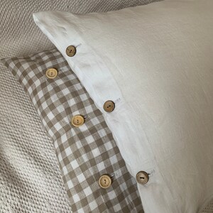 Puede incluir: Dos fundas de almohada decorativas. Una es blanca, la otra tiene un estampado de cuadros vichy marrón y blanco. Ambas tienen botones de madera en el borde. Las almohadas están hechas de una tela suave y texturizada.