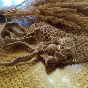 May include: A handmade, open-weave crochet bag in a natural brown color, featuring a decorative flower detail. The bag has a long strap and is displayed with dried ornamental grasses in a vase.
