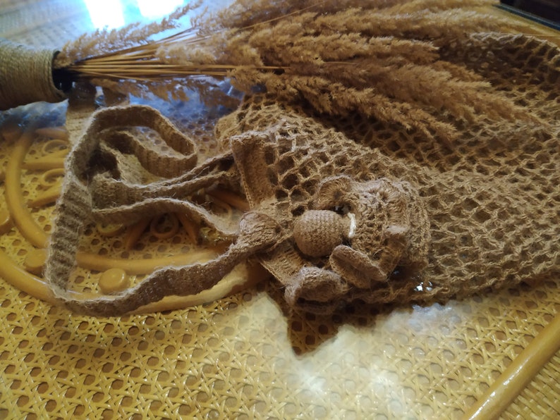 May include: A handmade, open-weave crochet bag in a natural brown color, featuring a decorative flower detail. The bag has a long strap and is displayed with dried ornamental grasses in a vase.