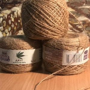 Peut inclure: Trois bobines de ficelle marron naturel sont empilées sur une surface en bois. La ficelle est étiquetée avec du texte et un graphisme de feuille de cannabis. Les bobines sont en jute et mesurent 190 mètres de long.