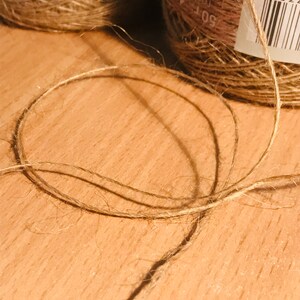 May include: Close-up of natural twine and spools of jute twine. The twine is a light brown color, with a slightly rough texture. The spools have labels with text in a foreign language. The twine is arranged in a circular shape on a wooden surface.