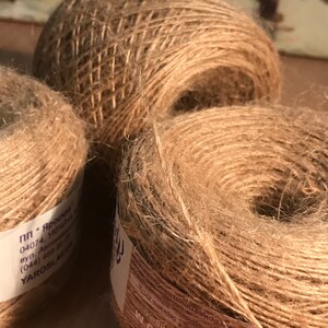 May include: Three spools of natural-colored twine, tightly wound and textured. The twine is a light brown color, with visible fibers. The spools are of varying sizes, with one larger spool in the background and two smaller ones in the foreground.