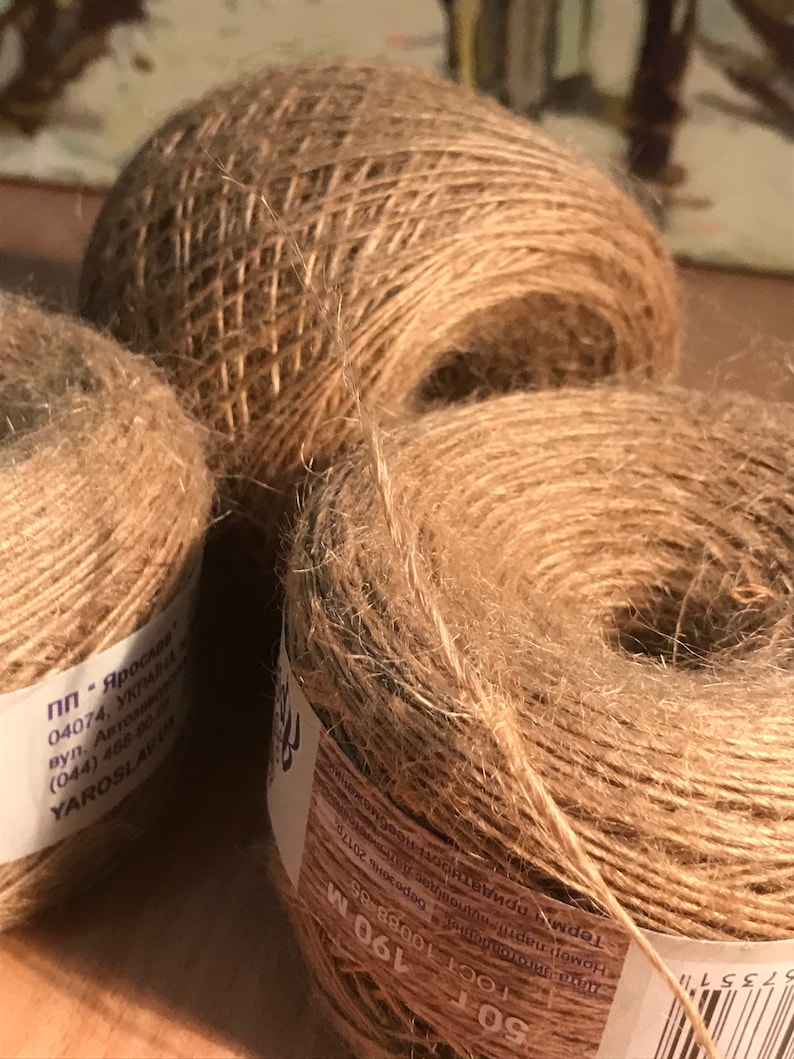 May include: Three spools of natural-colored twine, tightly wound and textured. The twine is a light brown color, with visible fibers. The spools are of varying sizes, with one larger spool in the background and two smaller ones in the foreground.