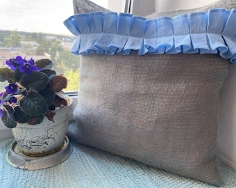 May include: A light blue pillow with a ruffled trim sits on a windowsill. The pillow is made of a burlap-like material and has a light brown color. A potted plant with purple flowers is in the foreground.