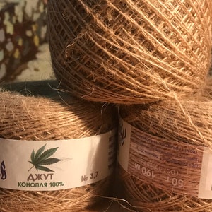 May include: Three balls of natural brown jute twine are stacked. Each ball is wrapped with a label that includes text in a foreign language and a cannabis leaf graphic. The twine is labeled "Jute Hemp 100%."