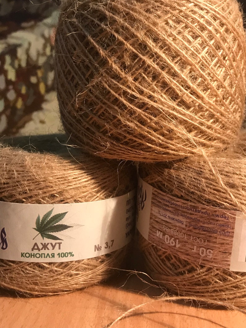 May include: Three balls of natural brown jute twine are stacked. Each ball is wrapped with a label that includes text in a foreign language and a cannabis leaf graphic. The twine is labeled "Jute Hemp 100%."