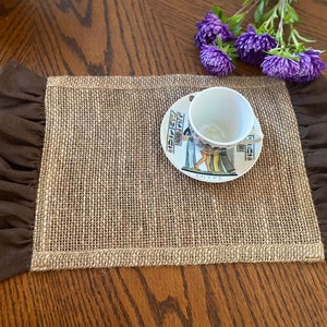 May include: A brown burlap placemat with a brown ruffled border. The placemat is set on a wooden table and has a white saucer with a teacup and a colourful design on it.