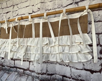 May include: A brown burlap valance with white ruffled trim. The valance is hung on a wooden rod and has white ribbon ties.