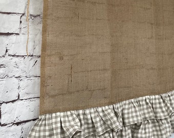 May include: A brown burlap window valance with a ruffled gingham trim. The valance is suspended from a wooden rod with twine. The background is a white brick wall.