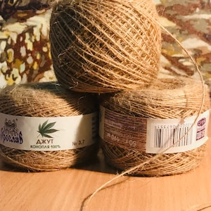 May include: Three spools of natural brown jute twine are stacked on a wooden surface. Each spool has a white label with text and a cannabis leaf graphic. The twine is a light brown color and appears to be made of natural fibers.