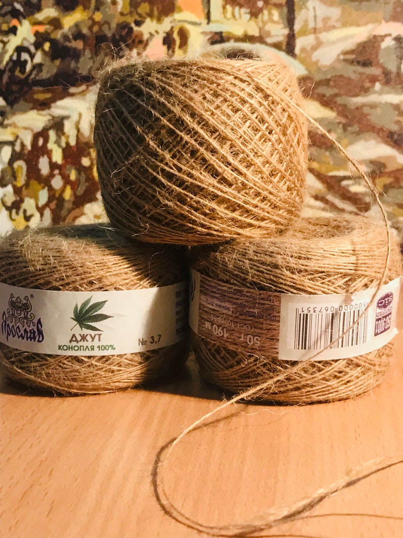 May include: Three spools of natural brown jute twine are stacked on a wooden surface. Each spool has a white label with text and a cannabis leaf graphic. The twine is a light brown color and appears to be made of natural fibers.
