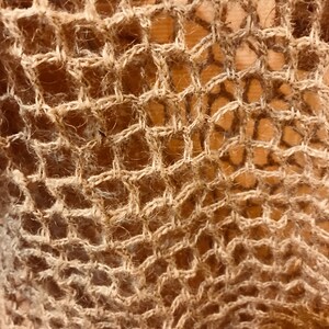May include: Close-up of a woven textile with a net-like pattern. The material is a light brown color, with a rough, textured appearance. The weave creates a series of diamond-shaped openings, giving the fabric an open, airy quality.