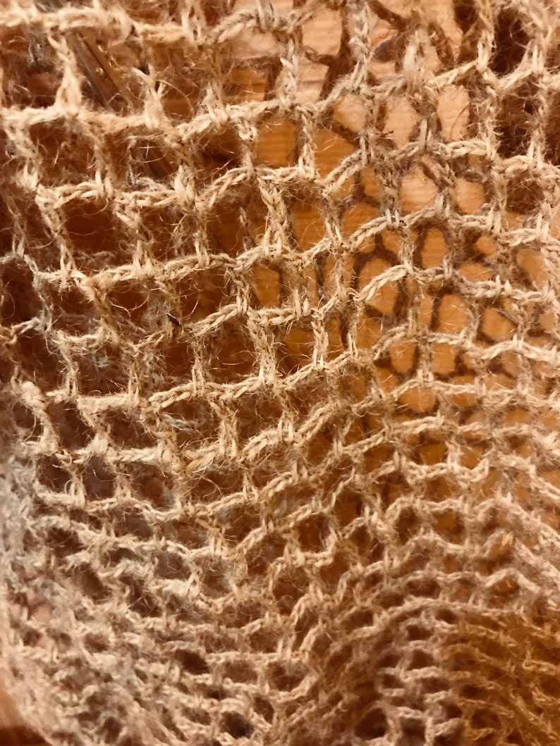 May include: Close-up of a woven textile with a net-like pattern. The material is a light brown color, with a rough, textured appearance. The weave creates a series of diamond-shaped openings, giving the fabric an open, airy quality.