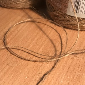May include: Close-up of natural jute twine, with a loop in the foreground. Two spools of jute twine are visible in the background. The twine is a light brown color, and the spools have labels with text.