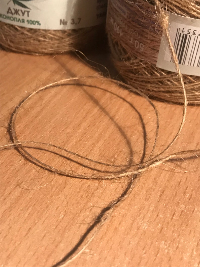 May include: Close-up of natural jute twine, with a loop in the foreground. Two spools of jute twine are visible in the background. The twine is a light brown color, and the spools have labels with text.
