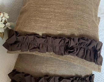 May include: Two brown burlap pillow covers with brown ruffled trim. The pillow covers are stacked on top of each other.