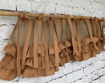 May include: A decorative valance with a rustic aesthetic. The valance features a burlap center panel, layered with a ruffled, terracotta-colored fabric. The valance is suspended from a wooden rod with matching fabric ties.