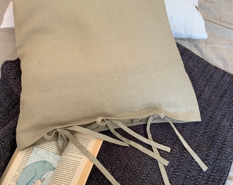 May include: A light green linen pillow with ties. The pillow is on a dark gray textured blanket with a book on top of it.