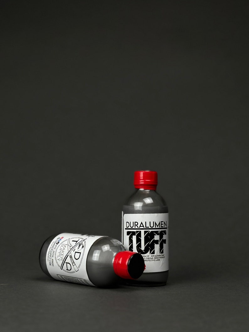 May include: Two small bottles of gray liquid with red caps. The bottle on the right has a white label with the text "Duralumen Tuff" and a description of the product. The bottle on the left has a white label with the text "TDD Maker Supply" and a description of the product.