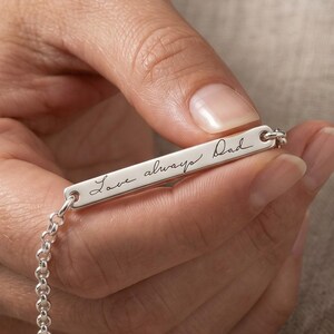 Peut inclure: Un bracelet en argent avec l'inscription "Love always Dad" est tenu dans une main. Le bracelet est orné d'une fine chaîne et est présenté sur un fond neutre.