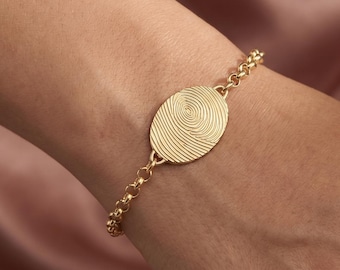 May include: A gold bracelet with an oval pendant engraved with a fingerprint pattern. The bracelet is attached to a delicate chain. The background is a soft, pinkish-peach color.