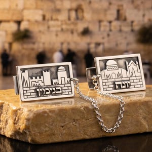 May include: Silver cufflinks with a rectangular design featuring architectural elements and Hebrew text. The cufflinks are connected by a small chain and rest on a light brown stone. The background shows a stone wall and blurred figures.