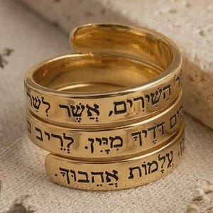 May include: A gold-colored wrap ring with three bands, each inscribed with black Hebrew text. The ring is coiled, with the bands overlapping. The background is a neutral beige surface with decorative elements.