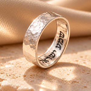 May include: A silver band ring featuring a hammered exterior texture and the Hebrew inscription Shema Yisrael engraved in black lettering along the inner surface of the metal band, creating a distinct contrast against the polished silver finish.