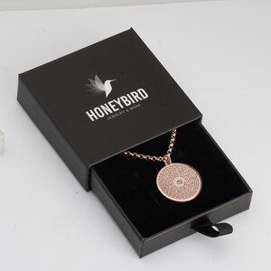 May include: A rose gold circular pendant necklace with a central stone and engraved text, presented in a black sliding jewellery box. The box lid features a white hummingbird logo and the text HONEYBIRD JEWELRY & MORE printed in white.