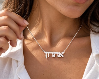 May include: A silver-toned necklace featuring a nameplate pendant with Hebrew characters. The pendant is suspended from a delicate box chain, resting against the collarbone. The metal has a polished, reflective finish that catches the light.