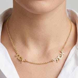 May include: A gold necklace featuring Hebrew script spelling two names. The necklace is worn against a white shirt. The chain is fine, and the letters are in a decorative font. The necklace is a piece of jewelry.