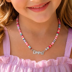 May include: A necklace featuring a string of small, multicolored, flat circular beads in shades of pink, blue, yellow, and white. A silver-toned metal nameplate with the name Emma is centered on the strand, attached by two small heart-shaped links.