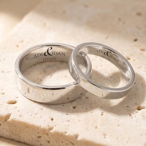 May include: Two polished silver-toned metal rings with flat bands. The inner surfaces are engraved with the text ADI & IDAN and the date 14.2.2025. The rings are positioned together, with the smaller ring partially overlapping the larger ring.