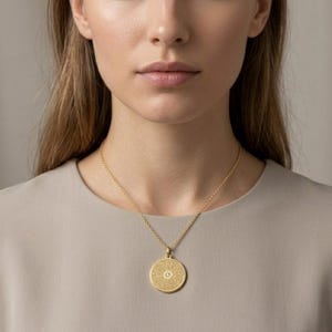 May include: A gold-toned necklace featuring a circular pendant with a textured surface and a small, centred clear stone. The pendant hangs from a delicate gold-toned chain, resting against a plain, light-coloured fabric garment.