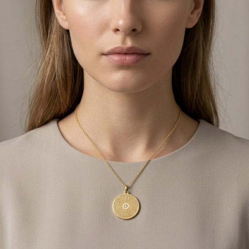May include: A gold-toned necklace featuring a circular pendant with a textured surface and a small, centred clear stone. The pendant hangs from a delicate gold-toned chain, resting against a plain, light-coloured fabric garment.