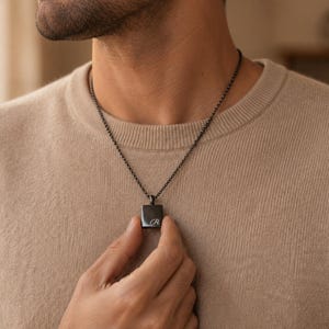May include: A black metal pendant necklace featuring a small, rectangular charm with the letter R engraved in a script font. The pendant hangs from a thin, matching black metal chain, resting against a beige knit sweater.