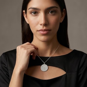 May include: A silver-toned circular pendant necklace featuring a textured, dotted surface pattern. The round medallion hangs from a delicate silver-toned chain, resting against a black garment with a cutout neckline detail.