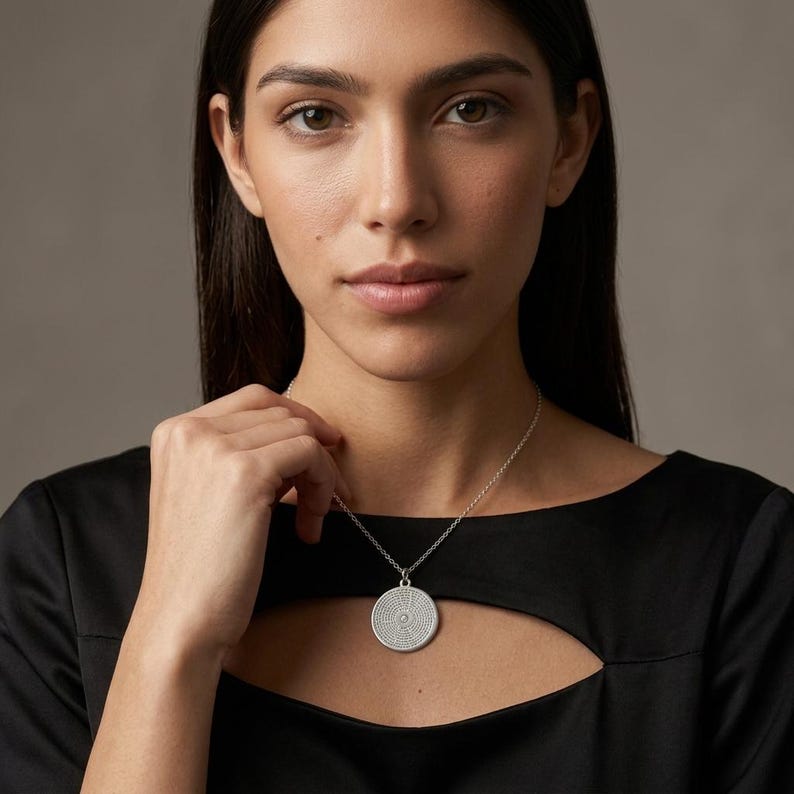 May include: A silver-toned circular pendant necklace featuring a textured, dotted surface pattern. The round medallion hangs from a delicate silver-toned chain, resting against a black garment with a cutout neckline detail.