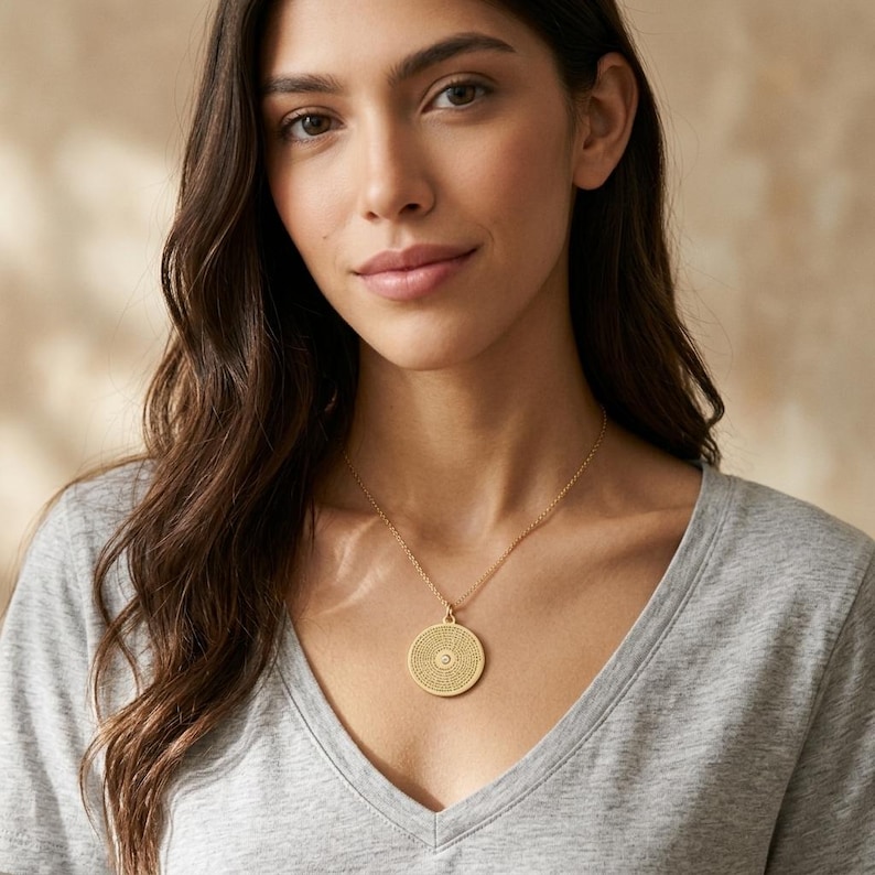 May include: A gold-toned circular pendant necklace featuring a textured, intricate pattern across the surface and a small, centred clear stone. The pendant hangs from a delicate gold-toned chain, resting against a plain grey v-neck shirt.