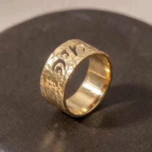 May include: A wide gold band ring featuring a hammered, textured surface and a series of cut-out, abstract, curved shapes along the upper edge. The metal has a polished, reflective finish that catches the light across its uneven, dimpled exterior.