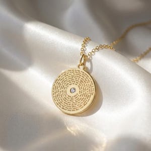 May include: A gold-toned circular pendant necklace featuring a central clear gemstone and intricate Hebrew text engraved across the entire surface. The pendant is suspended from a delicate matching gold-toned chain link necklace.